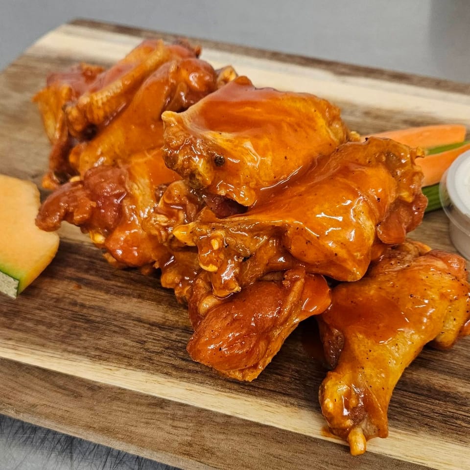Glazed chicken wings with carrots, celery, dipping sauce, and melon on a wooden board.
