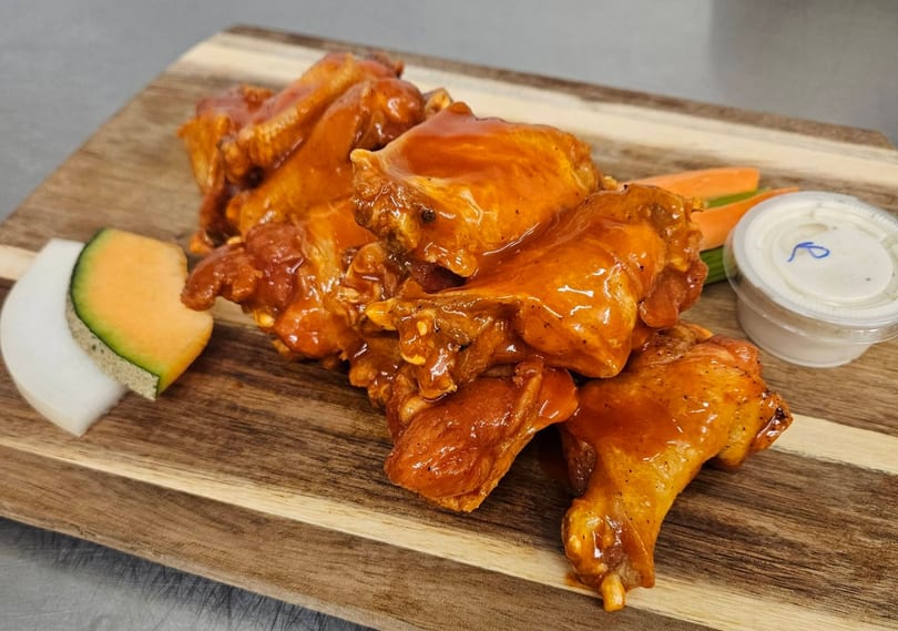 Glazed chicken wings with carrots, celery, dipping sauce, and melon on a wooden board.