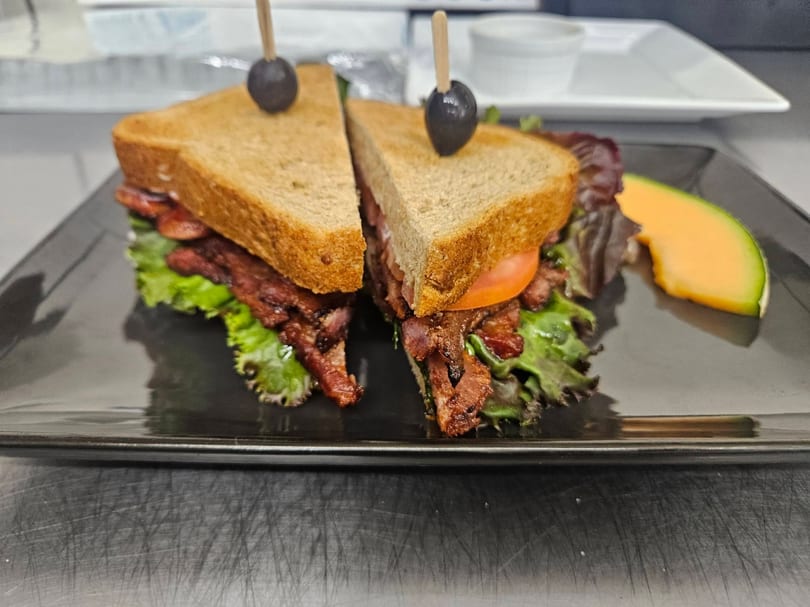 Toasted BLT sandwich cut in half, garnished with olives and a side of cantaloupe.