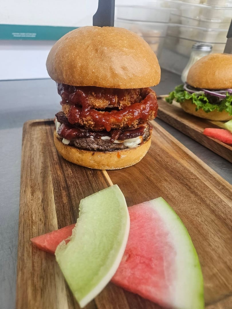 A tall burger with onion rings and BBQ sauce, served with watermelon and honeydew on a wooden board.