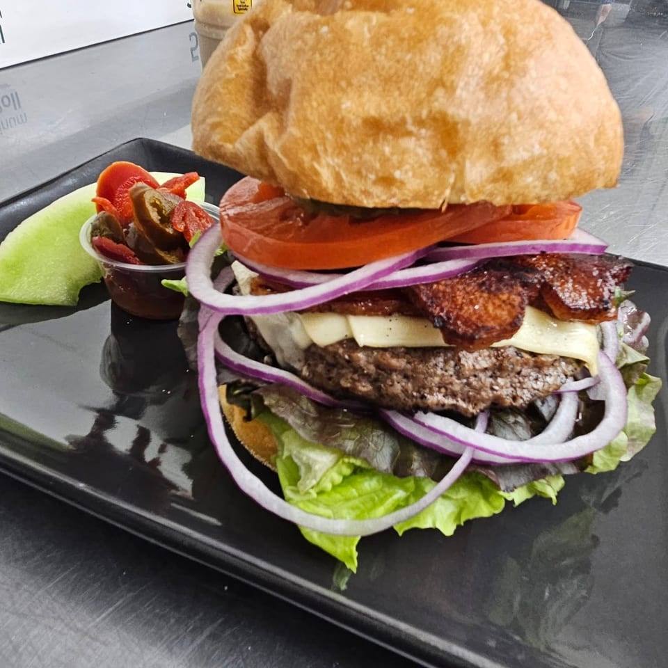 Gourmet burger with bacon, cheese, red onion, tomato, and lettuce, served with pickled peppers and melon.