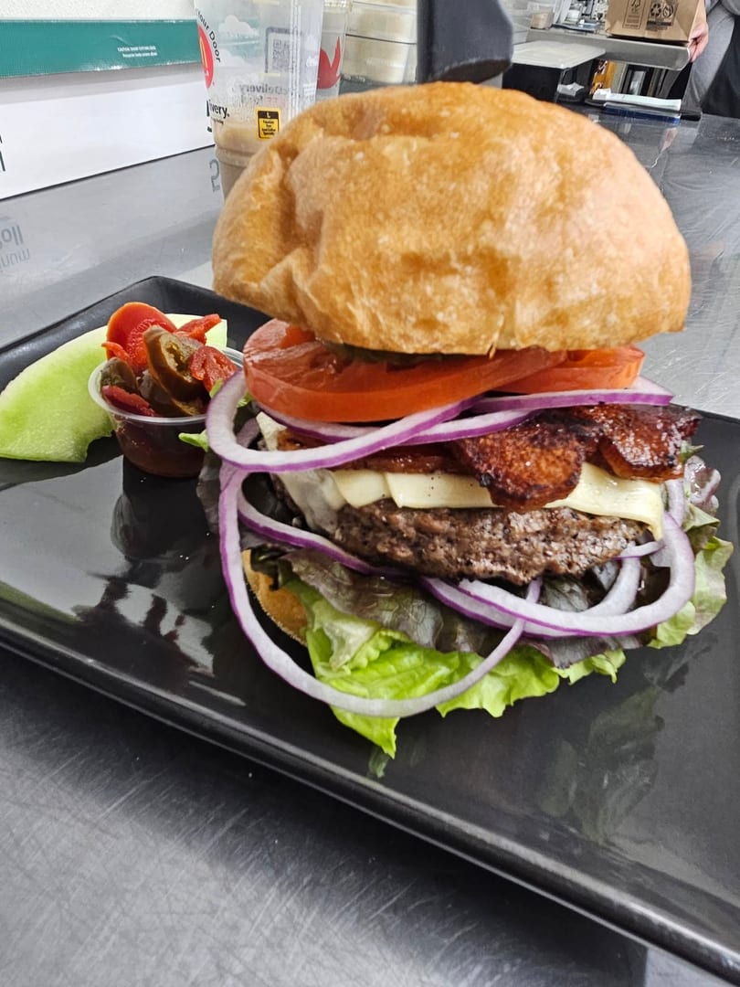 Gourmet burger with bacon, cheese, red onion, tomato, and lettuce, served with pickled peppers and melon.